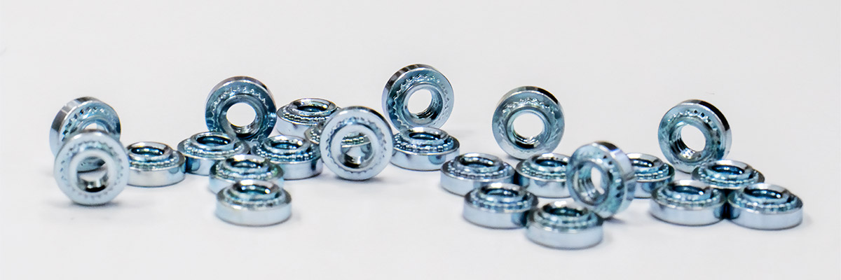 Vista en primer plano de diversos sujetadores, incluyendo tipos autoremachables, de brochado, de montaje en superficie y para soldar, dispuestos sobre una superficie metálica para mostrar diferentes formas y acabados.